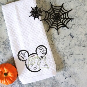 Może przedstawiać: Biały ręcznik z tkaniny o strukturze wafla z czarno-zieloną grafiką postaci i napisem "Let's Oogie Boogie". Na zdjęciu znajdują się r&oacute;wnież dwie czarne pajęczyny i pomarańczowa dynia. Motyw Halloween.