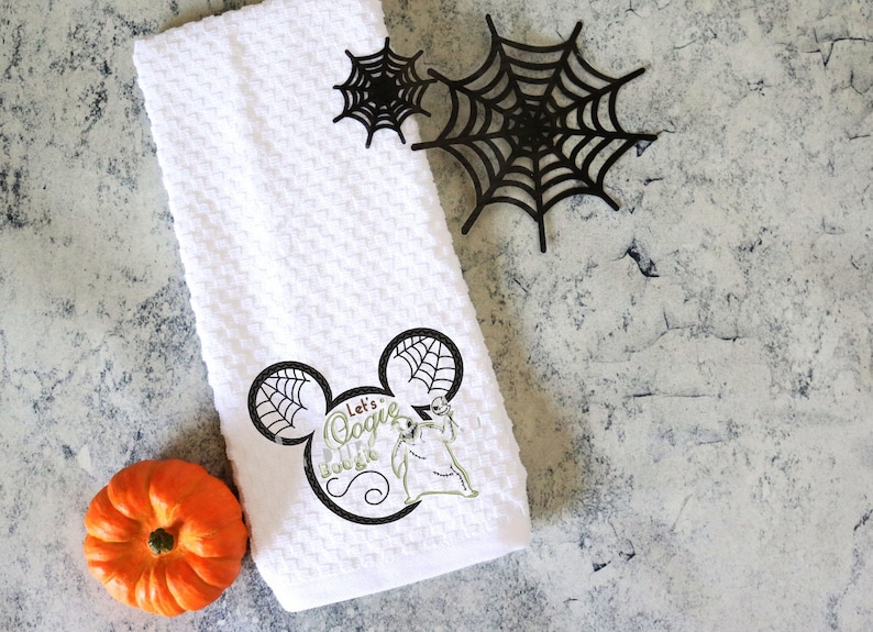 Może przedstawiać: Biały ręcznik z tkaniny o strukturze wafla z czarno-zieloną grafiką postaci i napisem "Let's Oogie Boogie". Na zdjęciu znajdują się r&oacute;wnież dwie czarne pajęczyny i pomarańczowa dynia. Motyw Halloween.
