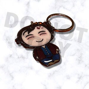 May include: A cartoon-style keychain featuring a smiling character with brown hair, a blue shirt, and a brown jacket. The keychain has a bronze-colored ring attached to the top. The character has a few red marks on their forehead.
