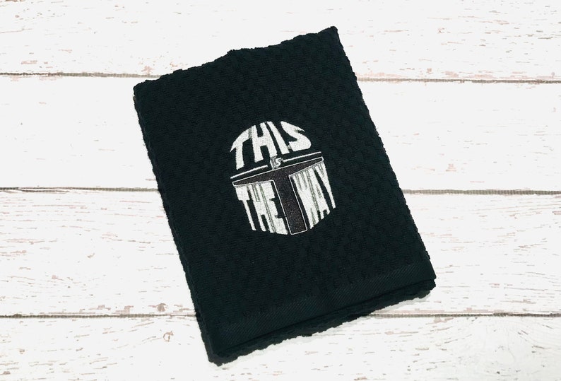 Puede incluir: Toalla de mano negra con una superficie texturizada. La toalla presenta un gr&aacute;fico blanco de un casco con el texto "THIS IS THE WAY" en una fuente estilizada. La toalla est&aacute; doblada y colocada sobre una superficie de madera blanca.