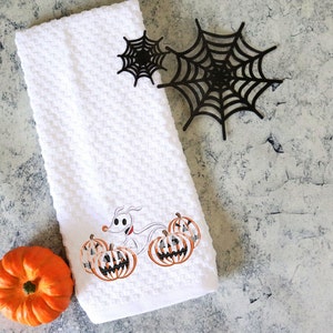 Może przedstawiać: Biały ręcznik kuchenny o tematyce Halloween z haftowanym wzorem przedstawiającym postać z kresk&oacute;wki psa i dynie. Zawiera małą pomarańczową dynię i czarne dekoracje z pajęczyny. Ręcznik ma wzorzysty, gofrowy wz&oacute;r.