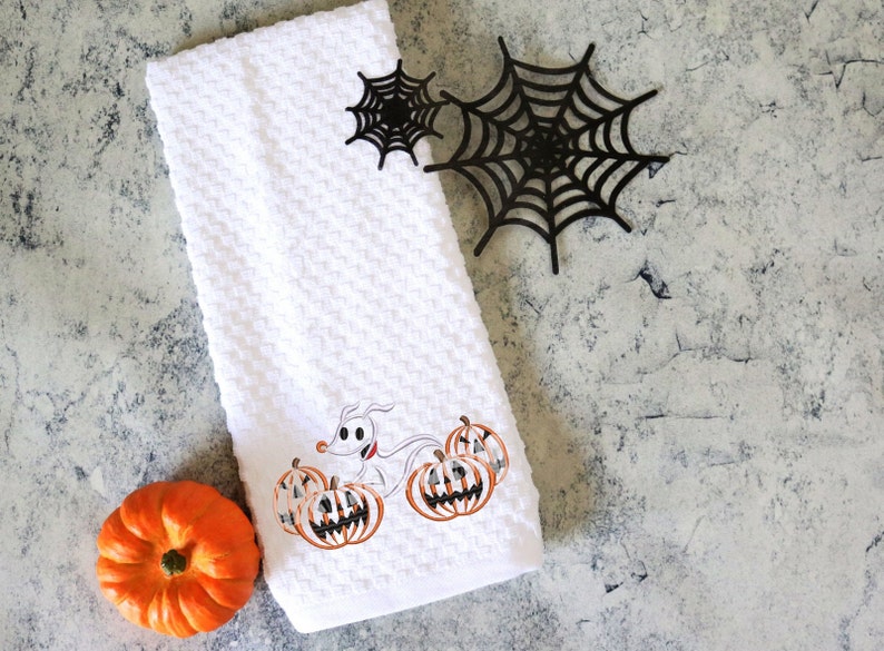 Może przedstawiać: Biały ręcznik kuchenny o tematyce Halloween z haftowanym wzorem przedstawiającym postać z kresk&oacute;wki psa i dynie. Zawiera małą pomarańczową dynię i czarne dekoracje z pajęczyny. Ręcznik ma wzorzysty, gofrowy wz&oacute;r.