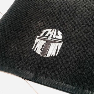 Puede incluir: Tela negra texturizada con un dise&ntilde;o bordado. El dise&ntilde;o es un casco estilizado con las palabras "THIS IS THE WAY" en blanco y gris. La tela tiene un patr&oacute;n tejido.