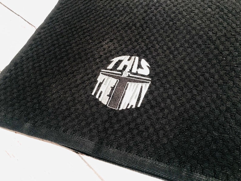 Puede incluir: Tela negra texturizada con un dise&ntilde;o bordado. El dise&ntilde;o es un casco estilizado con las palabras "THIS IS THE WAY" en blanco y gris. La tela tiene un patr&oacute;n tejido.