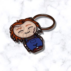 May include: A Chucky-themed keychain featuring a cartoon-style design. The keychain depicts Chucky with red hair, stitched scars, and blue overalls. The metal ring is a bronze color. The text "GOOD GUY" is visible on the character's face.