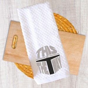 Puede incluir: Toalla de cocina blanca con tejido de gofre y la frase "THIS IS THE WAY" bordada en gris y negro. La toalla est&aacute; doblada y descansa sobre una tabla de cortar de madera y un salvamanteles tejido.