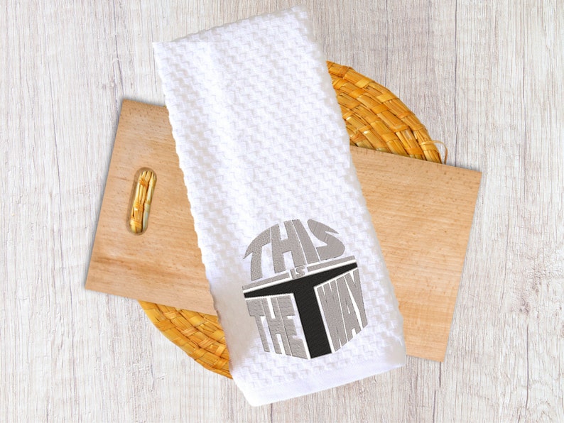 Puede incluir: Toalla de cocina blanca con tejido de gofre y la frase "THIS IS THE WAY" bordada en gris y negro. La toalla est&aacute; doblada y descansa sobre una tabla de cortar de madera y un salvamanteles tejido.
