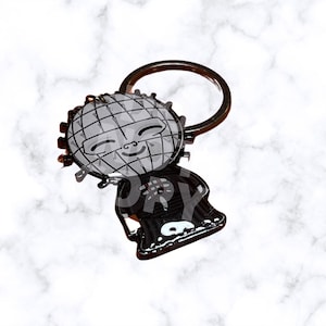 May include: A metal keychain featuring a cartoon character with a grid-like head and a smiling face. The character is wearing a black outfit with a red design. The keychain has a silver ring.