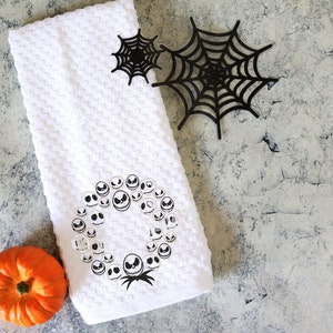 Może przedstawiać: Biały ręcznik o strukturze wafla z wieńcem z twarzy Jacka Skellingtona, małą pomarańczową dynią i dwiema czarnymi dekoracjami z pajęczyny. Ręcznik ma motyw Halloween.
