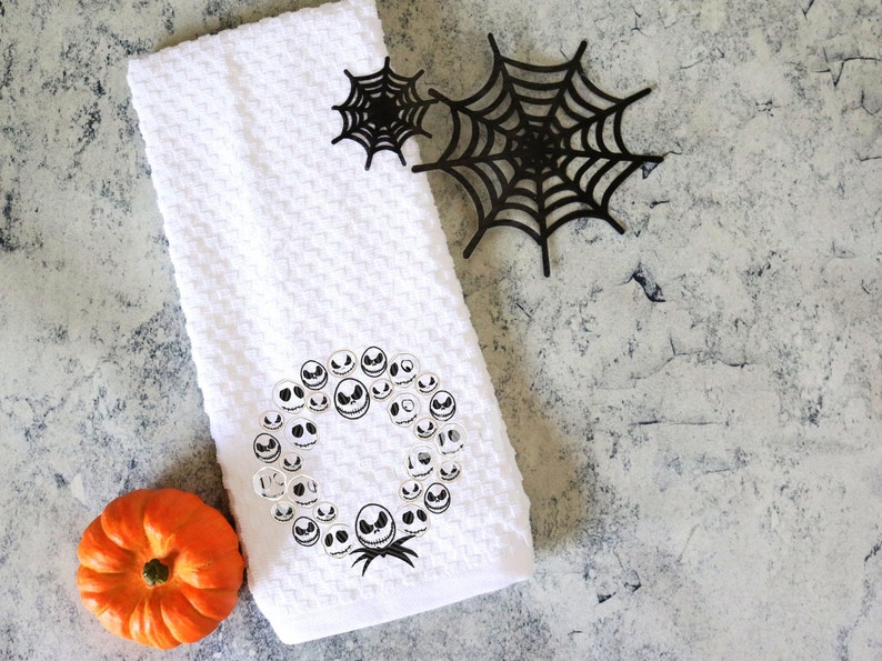 Może przedstawiać: Biały ręcznik o strukturze wafla z wieńcem z twarzy Jacka Skellingtona, małą pomarańczową dynią i dwiema czarnymi dekoracjami z pajęczyny. Ręcznik ma motyw Halloween.