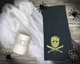 May include: A Halloween-themed image featuring a black hand towel with an embroidered Jason Voorhees mask and crossed machetes. A white ceramic mug with the word "GHOST" is next to a draped white fabric ghost. Black plastic spiders and a wooden sign that says "BOO" complete the spooky scene.