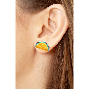 May include: A single taco-shaped earring is visible on an ear. The earring features a yellow tortilla, red tomatoes, and blue lettuce. The earring is a fun, food-themed accessory.