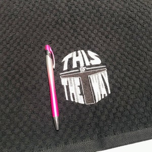 Puede incluir: Toalla de cocina negra con un patr&oacute;n texturizado. La frase "THIS IS THE WAY" est&aacute; impresa en blanco, junto con un dise&ntilde;o de casco. Un bol&iacute;grafo rosa y plateado descansa sobre la toalla.