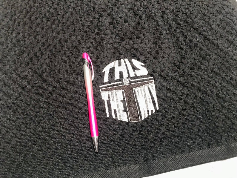 Puede incluir: Toalla de cocina negra con un patr&oacute;n texturizado. La frase "THIS IS THE WAY" est&aacute; impresa en blanco, junto con un dise&ntilde;o de casco. Un bol&iacute;grafo rosa y plateado descansa sobre la toalla.