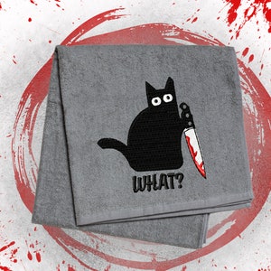 May include: Gray folded towel with an embroidered black cat holding a bloody knife. The word "WHAT?" is embroidered below the cat. The background has red paint splatters and a red circle.