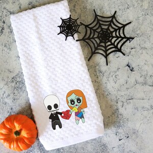Może przedstawiać: Biały ręcznik do rąk o strukturze wafla z kresk&oacute;wkową ilustracją szkieletu i zombie trzymających serce. Obecna jest r&oacute;wnież mała pomarańczowa dynia i czarne dekoracje z pajęczyny. Ręcznik dekoracyjny o tematyce Halloween.
