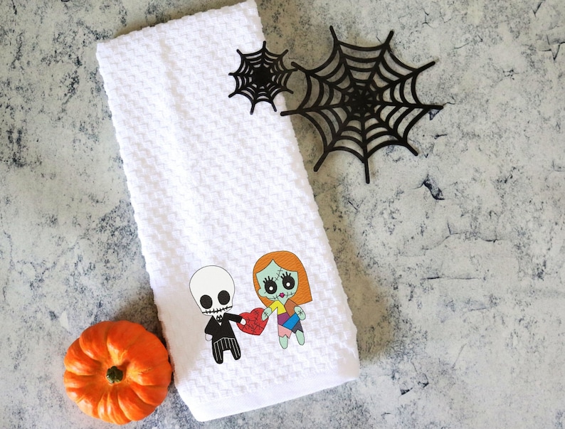 Może przedstawiać: Biały ręcznik do rąk o strukturze wafla z kresk&oacute;wkową ilustracją szkieletu i zombie trzymających serce. Obecna jest r&oacute;wnież mała pomarańczowa dynia i czarne dekoracje z pajęczyny. Ręcznik dekoracyjny o tematyce Halloween.