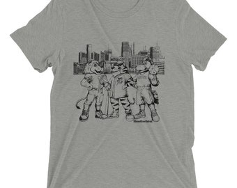 Puede incluir: Una camiseta gris con una ilustración en blanco y negro de tres mascotas de dibujos animados frente a un horizonte de ciudad. Las mascotas llevan uniformes deportivos y el horizonte incluye el texto "Motor City".