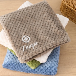 May include: A stack of four plush blankets in different colours, including brown, white, green, and blue. The top blanket is brown with a white compass design and the text "Dimple" embroidered on it.