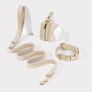 Könnte beinhalten: Ein beigefarbenes Hundehalsband, eine Leine und ein Kotbeutelhalter mit goldenen Beschlägen. Die Leine besteht aus einem weichen, strukturierten Material und hat einen goldenen Clip. Das Halsband besteht ebenfalls aus einem weichen, strukturierten Material und hat eine goldene Schnalle. Der Kotbeutelhalter ist eine kleine, rechteckige Tasche mit Reißverschluss und einem goldenen Clip.