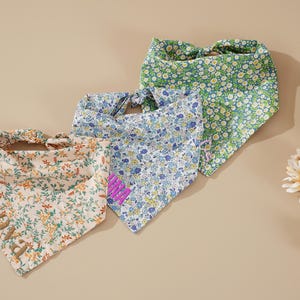 May include: Three floral patterned dog bandanas in cream, blue, and green. The cream bandana has the name "PRADA" embroidered in green. The blue bandana has a purple embroidered patch. The green bandana has a white floral pattern.