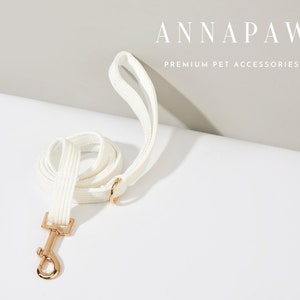 K&ouml;nnte beinhalten: Hunde-Leine aus wei&szlig;em Baumwollstoff mit einem goldenen Metallverschluss. Die Leine ist aufgerollt und der Markenname "ANNAPAW PREMIUM PET ACCESSORIES" ist oben im Bild zu sehen.