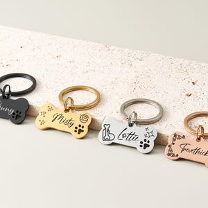 Pode incluir: Quatro etiquetas de identificação para animais de estimação em forma de osso em preto, dourado, prateado e ouro rosa. Cada etiqueta tem um nome diferente gravado, juntamente com pegadas e elementos decorativos.