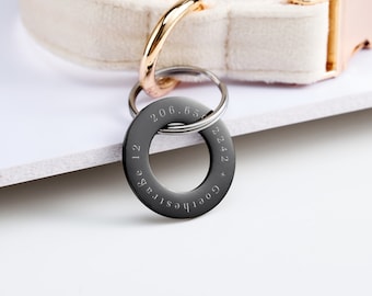 May include: A black, circular keychain with engraved text. The keychain is attached to a silver ring and a gold-colored ring. The text includes numbers and the words "Goethestrasse 12". The background is white and a beige collar is visible.