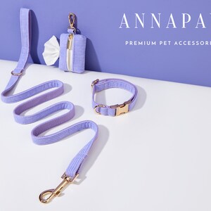 May include: A lavender pet leash, collar, and poop bag holder with gold hardware. The leash and collar are made of a soft, textured fabric. The poop bag holder has a zipper closure. The text "ANNAPAW PREMIUM PET ACCESSORIES" is displayed above the products.