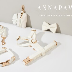 K&ouml;nnte beinhalten: Ein wei&szlig;es Hundegeschirr, eine Leine, ein Halsband und ein Kotbeutelhalter mit goldenen Beschl&auml;gen. Das Set ist auf einer wei&szlig;en Oberfl&auml;che angeordnet. Der Text "ANNAPAW PREMIUM PET ACCESSORIES" ist im Hintergrund sichtbar.