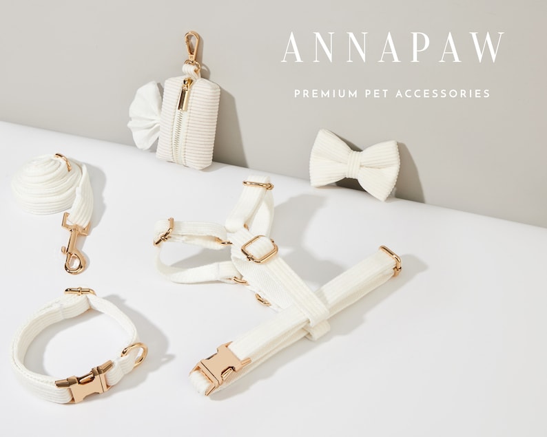 K&ouml;nnte beinhalten: Ein wei&szlig;es Hundegeschirr, eine Leine, ein Halsband und ein Kotbeutelhalter mit goldenen Beschl&auml;gen. Das Set ist auf einer wei&szlig;en Oberfl&auml;che angeordnet. Der Text "ANNAPAW PREMIUM PET ACCESSORIES" ist im Hintergrund sichtbar.