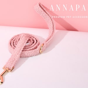 K&ouml;nnte beinhalten: Eine rosa flauschige Hundeleine mit einem goldenen Metallverschluss. Die Leine ist aufgerollt und hat den Text "ANNAPAW PREMIUM PET ACCESSORIES" auf rosa Hintergrund.
