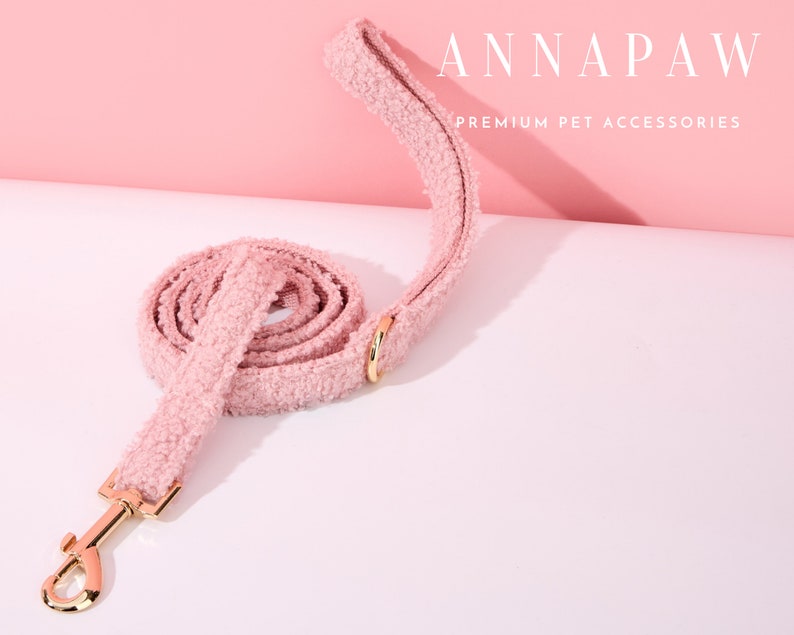 K&ouml;nnte beinhalten: Eine rosa flauschige Hundeleine mit einem goldenen Metallverschluss. Die Leine ist aufgerollt und hat den Text "ANNAPAW PREMIUM PET ACCESSORIES" auf rosa Hintergrund.