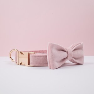 May include: A light pink dog collar and matching bow tie set. The collar has a gold-colored metal buckle and a D-ring. The bow tie is a classic shape. Both items are made of a soft, velvety material.