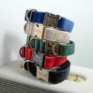 Pode incluir: Uma pilha de coleiras personalizadas para cães em várias cores, incluindo azul, bege, verde, vermelho e preto. Cada coleira apresenta uma fivela de metal gravada com um nome. As coleiras são feitas de um material macio, semelhante ao veludo.