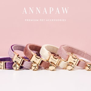Può includere: Cinque collari per gatti in velluto di diversi colori con targhette personalizzate e campanelle dorate. I collari sono in tonalità di viola, rosa e bianco. Il nome del marchio "ANNAPAW PREMIUM PET ACCESSORIES" è visualizzato nella parte superiore dell'immagine.