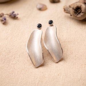 May include: A pair of silver-toned earrings with a curved, leaf-like design. Each earring features a small, dark-colored stone at the top. The earrings are displayed on a textured, beige surface, with natural elements in the background.
