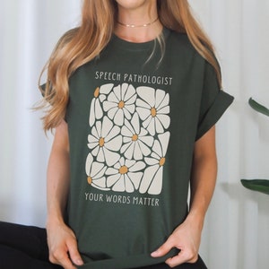 Puede incluir: Camiseta verde bosque con estampado de margaritas blancas y el texto "Speech Pathologist" y "Your Words Matter".
