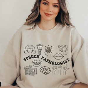 Puede incluir: Una sudadera beige con un dise&ntilde;o gr&aacute;fico negro que presenta ilustraciones anat&oacute;micas y el texto "SPEECH PATHOLOGIST".