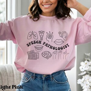 Puede incluir: Sudadera rosa claro con un dise&ntilde;o gr&aacute;fico de s&iacute;mbolos de pat&oacute;logo del habla, incluyendo un cerebro, pulmones, boca, o&iacute;do y manos de lenguaje de se&ntilde;as. El texto "Speech Pathologist" est&aacute; escrito en negro.