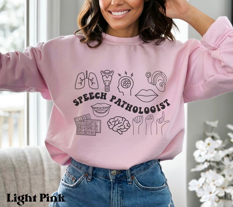 Puede incluir: Sudadera rosa claro con un dise&ntilde;o gr&aacute;fico de s&iacute;mbolos de pat&oacute;logo del habla, incluyendo un cerebro, pulmones, boca, o&iacute;do y manos de lenguaje de se&ntilde;as. El texto "Speech Pathologist" est&aacute; escrito en negro.