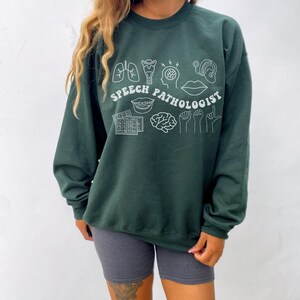 Puede incluir: Sudadera verde bosque con ilustraciones blancas de contorno de s&iacute;mbolos de pat&oacute;logo del habla y el texto "SPEECH PATHOLOGIST".