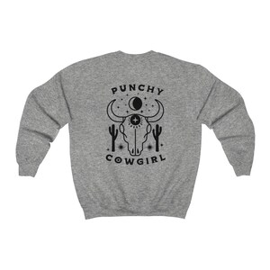 May include: Gray sweatshirt with a black graphic of a cow skull with a third eye, surrounded by stars and cacti. The text "Punchy Cowgirl" is below the skull.