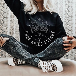 Puede incluir: Sudadera negra con un diseño de carnero blanco y el texto "HUGE ARIES ENERGY". Se combina con vaqueros desgastados y zapatillas con estampado de leopardo. El estilo es casual y moderno.