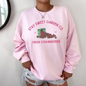 Puede incluir: Sudadera rosa con un gráfico de una caja llena de fresas y el texto "STAY SWEET GARDEN CO. EST 2021 FRESH STRAWBERRIES"