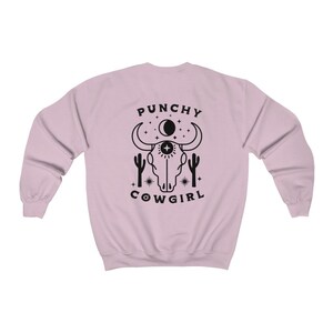 May include: A pink sweatshirt with a black graphic design of a cow skull with a third eye, surrounded by stars and cacti. The text "PUNCHY COWGIRL" is printed below the skull.