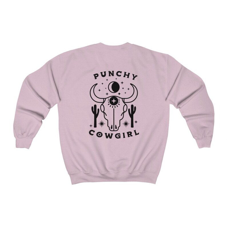 May include: A pink sweatshirt with a black graphic design of a cow skull with a third eye, surrounded by stars and cacti. The text "PUNCHY COWGIRL" is printed below the skull.