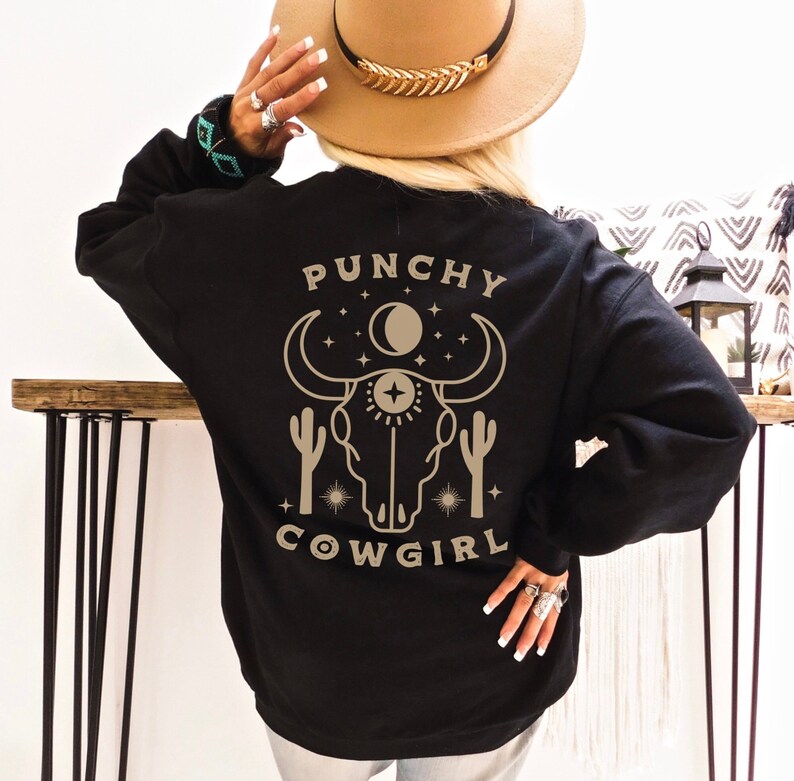 May include: Black sweatshirt with a graphic of a cow skull with a crescent moon and stars. The text "Punchy Cowgirl" is printed below the graphic.