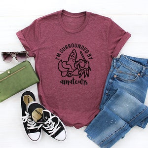 K&ouml;nnte beinhalten: Burgunderfarbenes T-Shirt mit dem Text "I'M SURROUNDED BY amateurs" in einem schwarzen, kreisf&ouml;rmigen Design. Das Bild enth&auml;lt auch eine gr&uuml;ne Geldb&ouml;rse, Sonnenbrille, schwarze Turnschuhe und blaue Jeans.