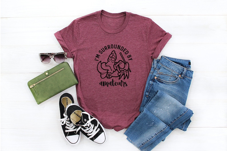 K&ouml;nnte beinhalten: Burgunderfarbenes T-Shirt mit dem Text "I'M SURROUNDED BY amateurs" in einem schwarzen, kreisf&ouml;rmigen Design. Das Bild enth&auml;lt auch eine gr&uuml;ne Geldb&ouml;rse, Sonnenbrille, schwarze Turnschuhe und blaue Jeans.